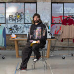 Australian Street Fashion Designer Embraces Generative AI with Caution A man with long hair and a beard, sitting on a chair in what seems like a studio or a store with clothes hanging on racks behind him