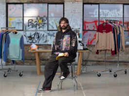 Australian Street Fashion Designer Embraces Generative AI with Caution A man with long hair and a beard, sitting on a chair in what seems like a studio or a store with clothes hanging on racks behind him