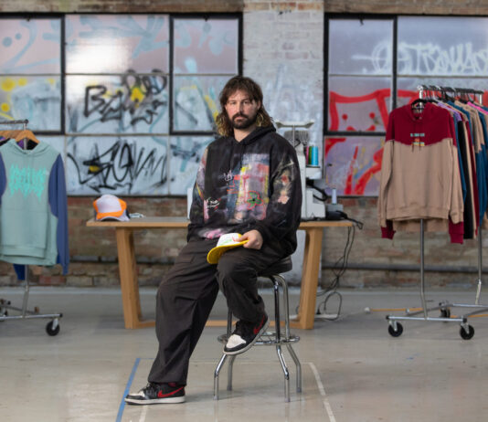 Australian Street Fashion Designer Embraces Generative AI with Caution A man with long hair and a beard, sitting on a chair in what seems like a studio or a store with clothes hanging on racks behind him