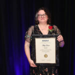 Misty Davies Named 2024 AIAA Fellow by NASA Misty Davies holding a framed AIAA Fellowship certificate with a red rose on her lapel, standing against a dark background.