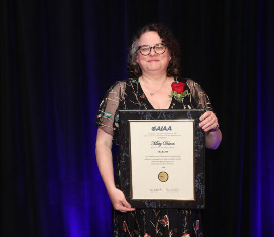 Misty Davies Named 2024 AIAA Fellow by NASA Misty Davies holding a framed AIAA Fellowship certificate with a red rose on her lapel, standing against a dark background.