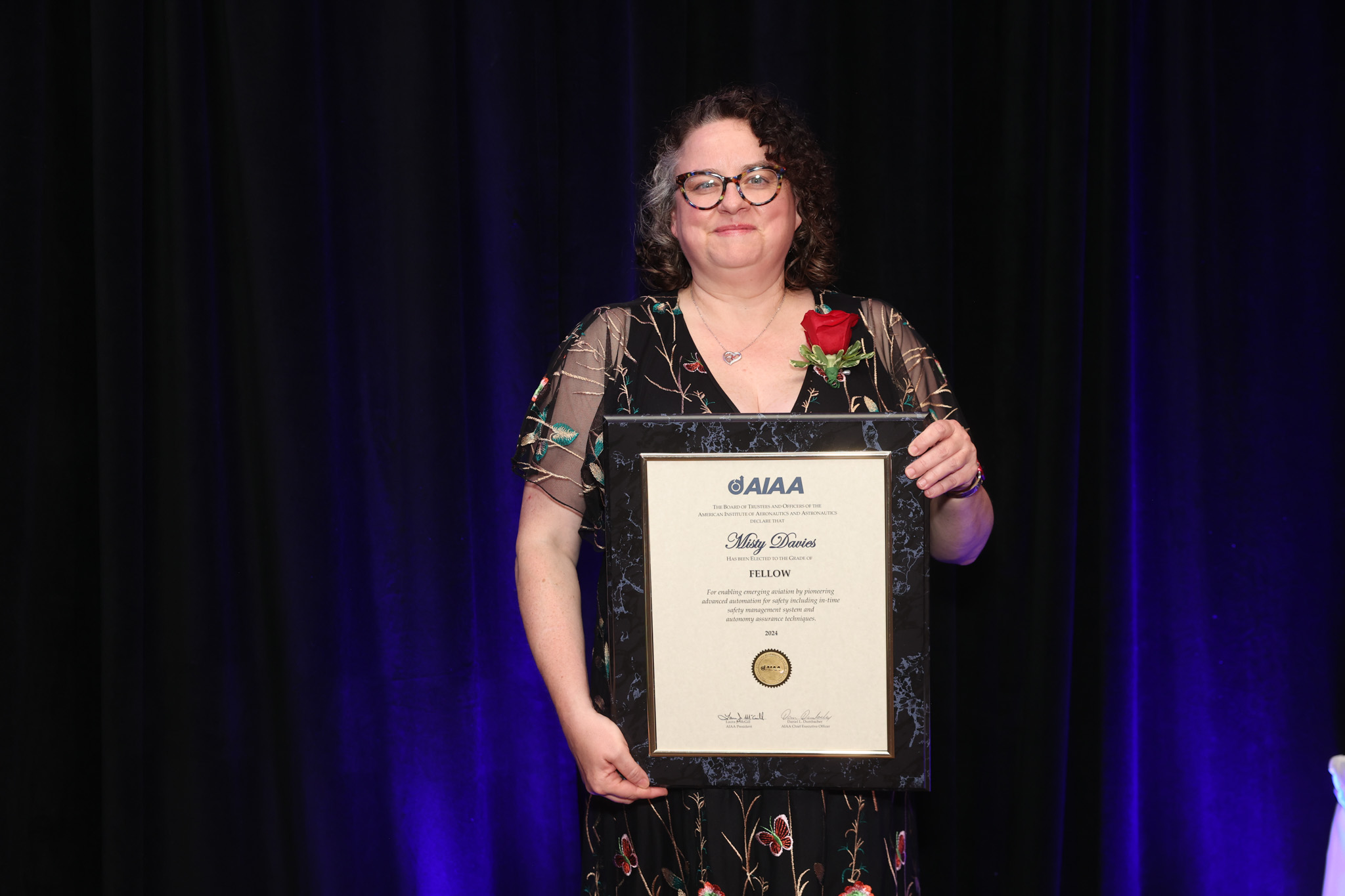 Misty Davies holding a framed AIAA Fellowship certificate with a red rose on her lapel, standing against a dark background.
