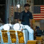 Starpath’s Break the Ice Winner Trials Robot in NASA Chamber A person wearing glasses and a dark polo shirt stands indoors holding an open laptop in one hand. In front of him is a piece of large mechanical equipment resembling a planetary surface rover, with white structural arms and panels covered in gold foil. Behind him, another person is carrying a large, flat, rectangular object. The background shows industrial equipment and a large American flag hanging on the wall.