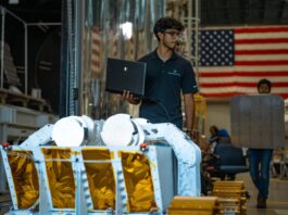 Starpath’s Break the Ice Winner Trials Robot in NASA Chamber A person wearing glasses and a dark polo shirt stands indoors holding an open laptop in one hand. In front of him is a piece of large mechanical equipment resembling a planetary surface rover, with white structural arms and panels covered in gold foil. Behind him, another person is carrying a large, flat, rectangular object. The background shows industrial equipment and a large American flag hanging on the wall.