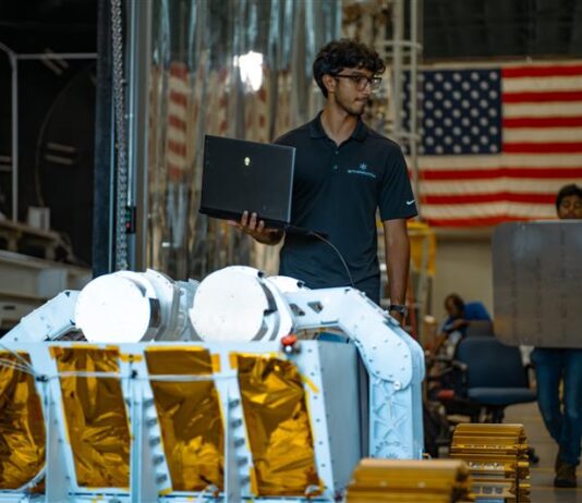 Starpath’s Break the Ice Winner Trials Robot in NASA Chamber A person wearing glasses and a dark polo shirt stands indoors holding an open laptop in one hand. In front of him is a piece of large mechanical equipment resembling a planetary surface rover, with white structural arms and panels covered in gold foil. Behind him, another person is carrying a large, flat, rectangular object. The background shows industrial equipment and a large American flag hanging on the wall.
