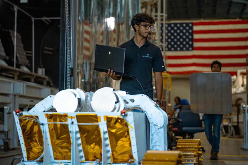 A person wearing glasses and a dark polo shirt stands indoors holding an open laptop in one hand. In front of him is a piece of large mechanical equipment resembling a planetary surface rover, with white structural arms and panels covered in gold foil. Behind him, another person is carrying a large, flat, rectangular object. The background shows industrial equipment and a large American flag hanging on the wall.