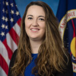 NASA’s Symphony of Simulations Crafted by Paige Whittington A white woman with long brown hair, wearing a blue blouse and black cardigan, sits in front of an American flag and a NASA flag.