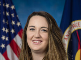 NASA’s Symphony of Simulations Crafted by Paige Whittington A white woman with long brown hair, wearing a blue blouse and black cardigan, sits in front of an American flag and a NASA flag.