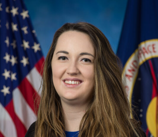 NASA’s Symphony of Simulations Crafted by Paige Whittington A white woman with long brown hair, wearing a blue blouse and black cardigan, sits in front of an American flag and a NASA flag.