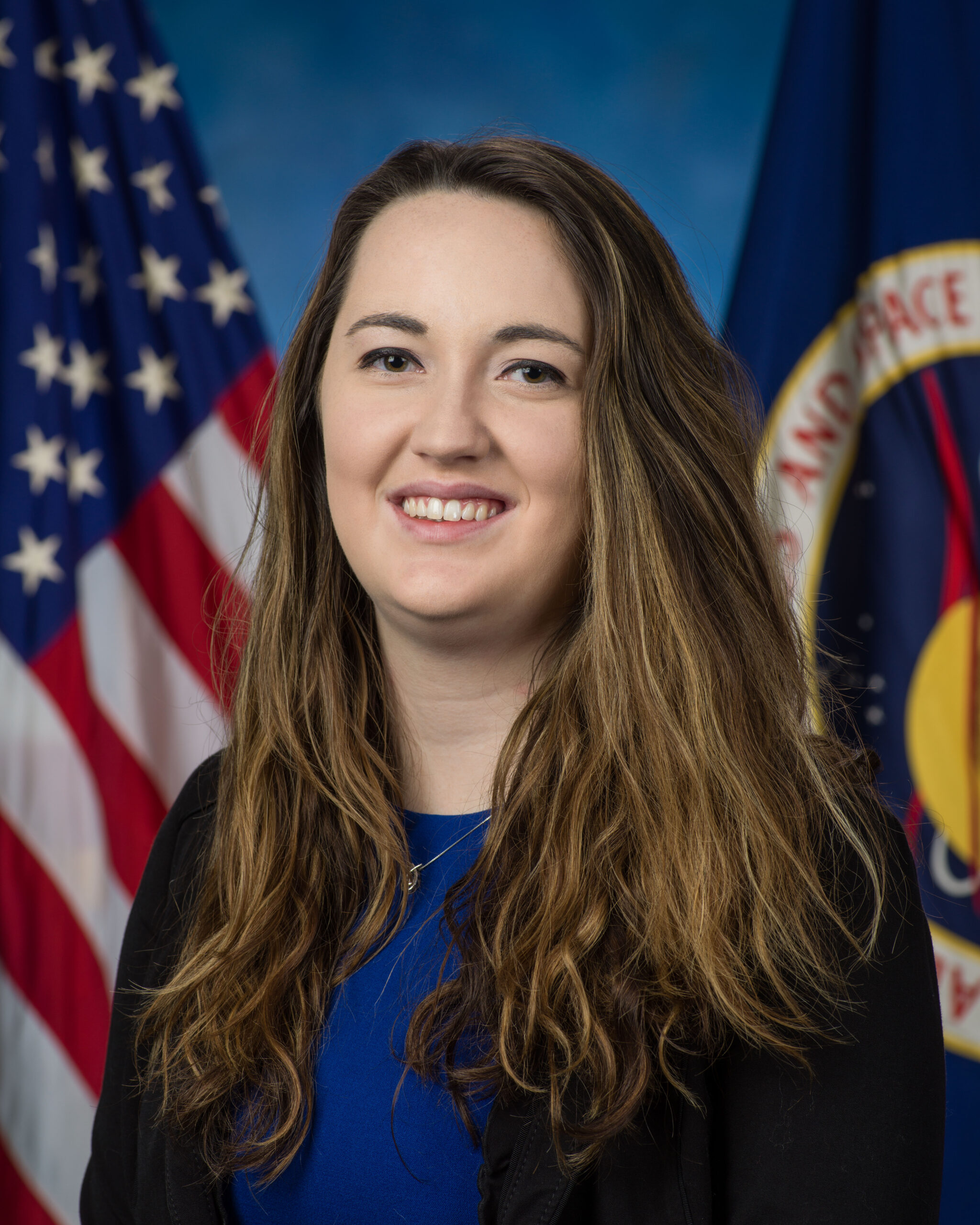 A white woman with long brown hair, wearing a blue blouse and black cardigan, sits in front of an American flag and a NASA flag.