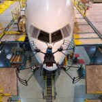 NASA Funds Drone Technology for Aircraft Inspection A small, black drone with four rotors is shown in the foreground flying in front of a commercial airliner in the background. The airliner is painted white and the front facing windows can be seen behind the drone. Concrete platforms surround the commercial airliner and yellow ramps connect the platforms to the plane.