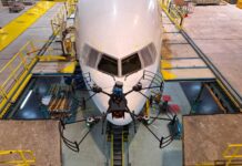 NASA Funds Drone Technology for Aircraft Inspection A small, black drone with four rotors is shown in the foreground flying in front of a commercial airliner in the background. The airliner is painted white and the front facing windows can be seen behind the drone. Concrete platforms surround the commercial airliner and yellow ramps connect the platforms to the plane.