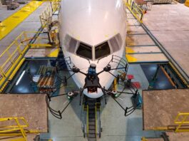 NASA Funds Drone Technology for Aircraft Inspection A small, black drone with four rotors is shown in the foreground flying in front of a commercial airliner in the background. The airliner is painted white and the front facing windows can be seen behind the drone. Concrete platforms surround the commercial airliner and yellow ramps connect the platforms to the plane.