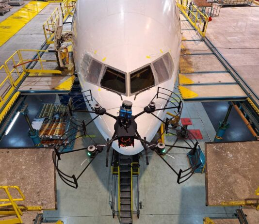 NASA Funds Drone Technology for Aircraft Inspection A small, black drone with four rotors is shown in the foreground flying in front of a commercial airliner in the background. The airliner is painted white and the front facing windows can be seen behind the drone. Concrete platforms surround the commercial airliner and yellow ramps connect the platforms to the plane.