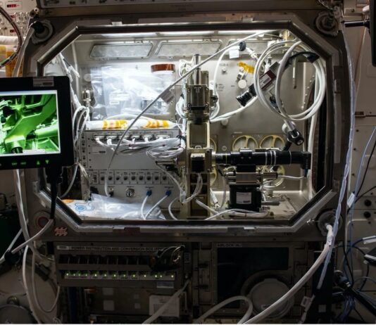 Adam and Hirsa Unveil NASA’s Ring-Sheared Drop Study A silver laboratory structure with wires and controls, connected to two laptops