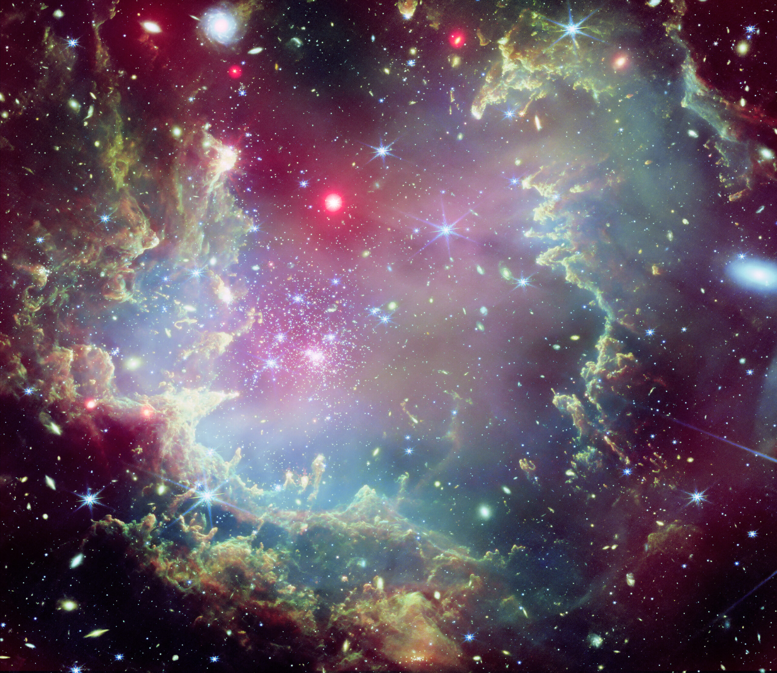 Near the outskirts of the Small Magellanic Cloud, a satellite galaxy roughly 200 000 light-years from Earth, lies the young star cluster NGC 602, which is featured in this new image from the NASA/ESA/CSA James Webb Space Telescope. This image includes data from Webb’s NIRCam (Near-InfraRed Camera) and MIRI (Mid-InfraRed Instrument). The local environment of this cluster is a close analogue of what existed in the early Universe, with very low abundances of elements heavier than hydrogen and helium. The existence of dark clouds of dense dust and the fact that the cluster is rich in ionised gas also suggest the presence of ongoing star formation processes. This cluster provides a valuable opportunity to examine star formation scenarios under dramatically different conditions from those in the solar neighbourhood. [Image description: A star cluster is shown inside a large nebula of many-coloured gas and dust. The material forms dark ridges and peaks of gas and dust surrounding the cluster, lit on the inner side, while layers of diffuse, translucent clouds blanket over them. Around and within the gas, a huge number of distant galaxies can be seen, some quite large, as well as a few stars nearer to us which are very large and bright.]