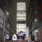 Celebrating Five Years Since SpaceX Demo-2 Launch with NASA A seated crowd is in the foreground, facing a stage in the distance where President Donald Trump stands at a podium. He is flanked by a SpaceX Dragon Crew capsule and part of a SpaceX Dragon rocket. The picture is taken inside of the Vehicle Assembly Building; scaffolding can be seen as you look up.