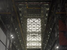 Celebrating Five Years Since SpaceX Demo-2 Launch with NASA A seated crowd is in the foreground, facing a stage in the distance where President Donald Trump stands at a podium. He is flanked by a SpaceX Dragon Crew capsule and part of a SpaceX Dragon rocket. The picture is taken inside of the Vehicle Assembly Building; scaffolding can be seen as you look up.