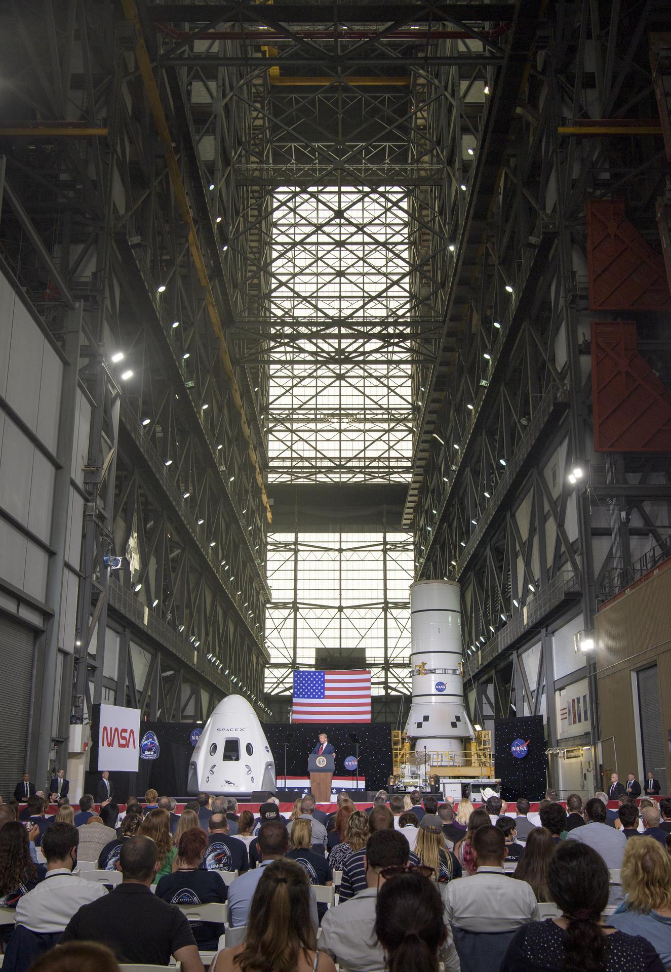 A seated crowd is in the foreground, facing a stage in the distance where President Donald Trump stands at a podium. He is flanked by a SpaceX Dragon Crew capsule and part of a SpaceX Dragon rocket. The picture is taken inside of the Vehicle Assembly Building; scaffolding can be seen as you look up.