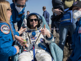 Astronauts Share Post-Microgravity Recovery Experiences Three people in blue flight suits stand around a woman in a white space suit who