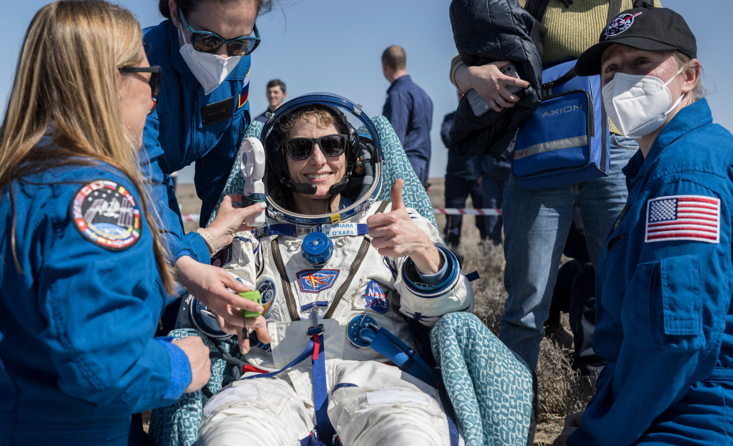 Three people in blue flight suits stand around a woman in a white space suit who