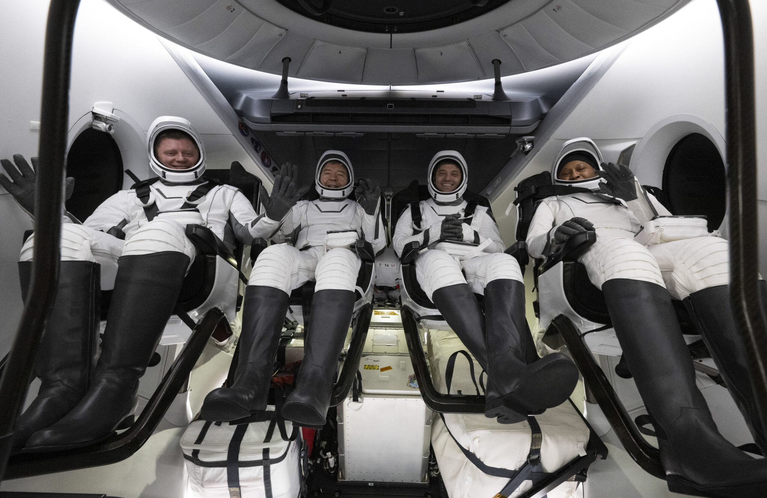 The four NASA SpaceX Crew-8 members sit in a row, smiling and waving in their spacesuits aboard a Dragon spacecraft.