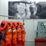 NASA reveals Artemis II Crew’s advanced space suits. Four neon orange spacesuits hang on a rack decorated with NASA patches. Three of the suits have American flag patches on their left shoulder, while the last has a Canadian flag patch on its left shoulder. The room the suits are hanging in has a large black and white photo on the wall, in which astronauts are in their spacesuits and helmets while two people in white lab coats look on.