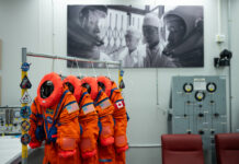 NASA reveals Artemis II Crew’s advanced space suits. Four neon orange spacesuits hang on a rack decorated with NASA patches. Three of the suits have American flag patches on their left shoulder, while the last has a Canadian flag patch on its left shoulder. The room the suits are hanging in has a large black and white photo on the wall, in which astronauts are in their spacesuits and helmets while two people in white lab coats look on.