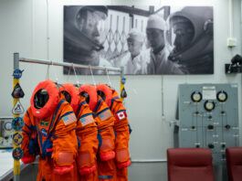 NASA reveals Artemis II Crew’s advanced space suits. Four neon orange spacesuits hang on a rack decorated with NASA patches. Three of the suits have American flag patches on their left shoulder, while the last has a Canadian flag patch on its left shoulder. The room the suits are hanging in has a large black and white photo on the wall, in which astronauts are in their spacesuits and helmets while two people in white lab coats look on.