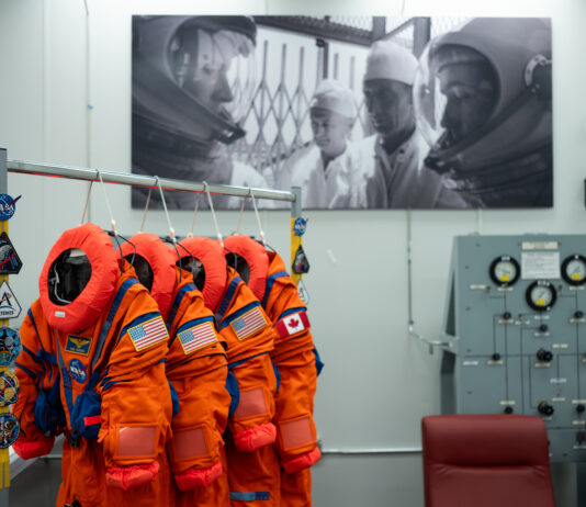 NASA reveals Artemis II Crew’s advanced space suits. Four neon orange spacesuits hang on a rack decorated with NASA patches. Three of the suits have American flag patches on their left shoulder, while the last has a Canadian flag patch on its left shoulder. The room the suits are hanging in has a large black and white photo on the wall, in which astronauts are in their spacesuits and helmets while two people in white lab coats look on.