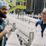 Obeikan’s AI Journey: Overcoming Challenges, Achieving Success Two men in blue hairnets speaking across a line of white plastic bottles on a conveyor belt.