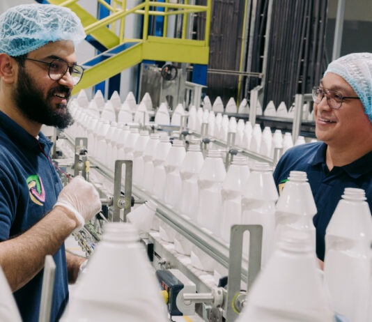 Obeikan’s AI Journey: Overcoming Challenges, Achieving Success Two men in blue hairnets speaking across a line of white plastic bottles on a conveyor belt.