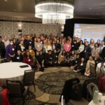 Amateur Scientists Explore Space Life with NASA’s Open Data Large group photo of attendees at a NASA Analysis Working Group meeting, gathered in a conference room with round tables and chandeliers. A presentation screen is visible in the background.