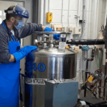 Exploring Careers: NASA’s Innovative Creation of ‘Memory Metals’ A man wearing a surgical mask, protective plastic face covering, and thick blue gloves, works with a large metal can that has the text