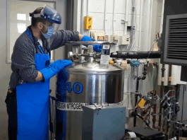 Exploring Careers: NASA’s Innovative Creation of ‘Memory Metals’ A man wearing a surgical mask, protective plastic face covering, and thick blue gloves, works with a large metal can that has the text