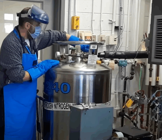 Exploring Careers: NASA’s Innovative Creation of ‘Memory Metals’ A man wearing a surgical mask, protective plastic face covering, and thick blue gloves, works with a large metal can that has the text