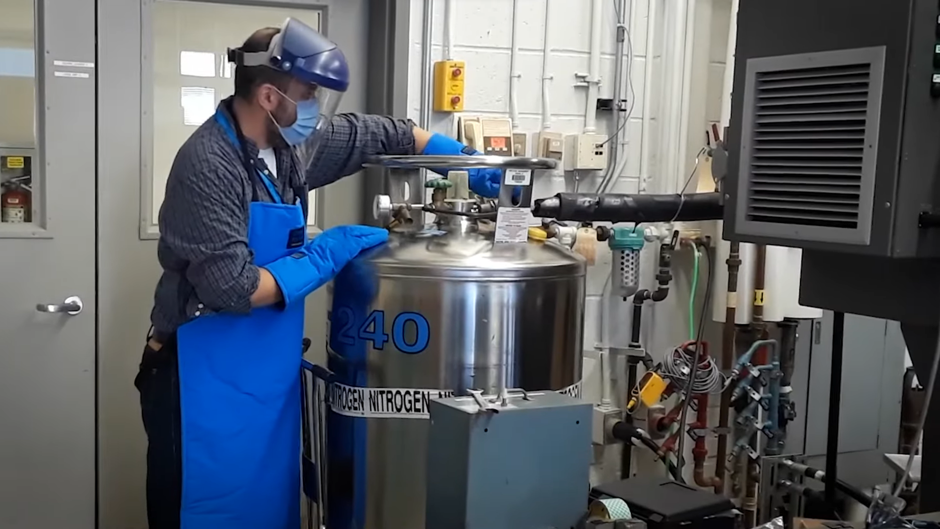 othmanebenafansurprisinglystem.png A man wearing a surgical mask, protective plastic face covering, and thick blue gloves, works with a large metal can that has the text