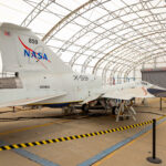 NASA X-59 Achieves Ground-Based Flight Simulation Milestone A white NASA jet airplane sits inside a fabric-covered-roof hangar with several people working at a nearby table with computers.