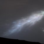 NASA Captures Rainbow-Hued “Feather” Cloud in Mars’ Atmosphere A shining, feather-shaped cloud in the dark gray Martian sky. The cloud is white, with a faint rainbow tint.