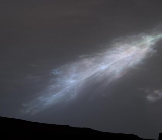 NASA Captures Rainbow-Hued “Feather” Cloud in Mars’ Atmosphere A shining, feather-shaped cloud in the dark gray Martian sky. The cloud is white, with a faint rainbow tint.