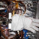 NASA Mobilizes Team for Artemis III Mission A bulky white spacesuit glove with gray fingertips sits inside a small chamber lined with silvery material. Multiple wires wrap around the fingers of the glove and loop around the floor of the chamber beneath the glove.