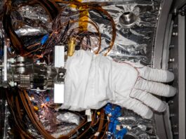 NASA Mobilizes Team for Artemis III Mission A bulky white spacesuit glove with gray fingertips sits inside a small chamber lined with silvery material. Multiple wires wrap around the fingers of the glove and loop around the floor of the chamber beneath the glove.