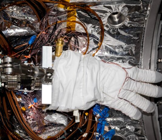 NASA Mobilizes Team for Artemis III Mission A bulky white spacesuit glove with gray fingertips sits inside a small chamber lined with silvery material. Multiple wires wrap around the fingers of the glove and loop around the floor of the chamber beneath the glove.