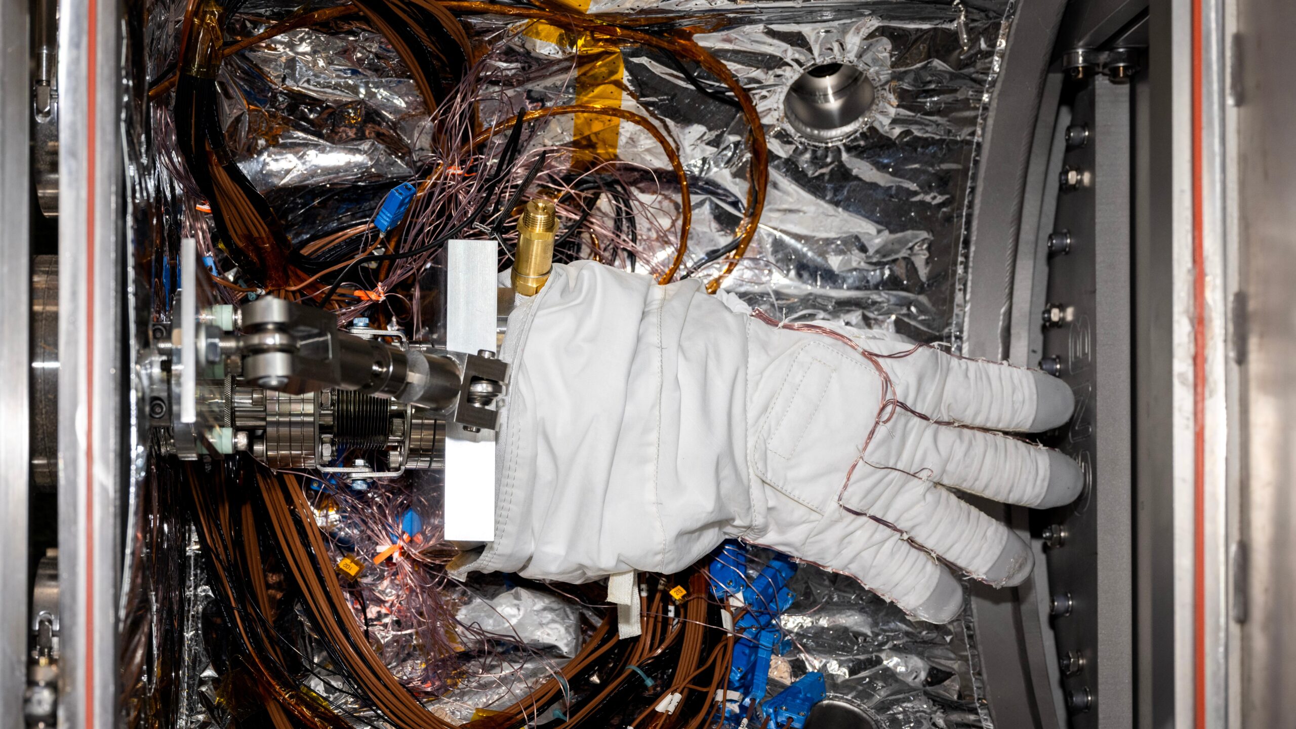 pia26430.jpg A bulky white spacesuit glove with gray fingertips sits inside a small chamber lined with silvery material. Multiple wires wrap around the fingers of the glove and loop around the floor of the chamber beneath the glove.