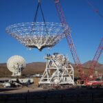 NASA Constructs New Antenna A large red crane lowers a cone-shaped frame. There is a large white metal structure behind it that has stairs running around it, all the way to the top. In the background at left, a completed antenna faces away from us. Farther away in the background are brown mountains.