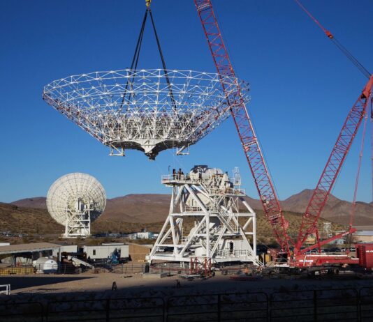 NASA Constructs New Antenna A large red crane lowers a cone-shaped frame. There is a large white metal structure behind it that has stairs running around it, all the way to the top. In the background at left, a completed antenna faces away from us. Farther away in the background are brown mountains.