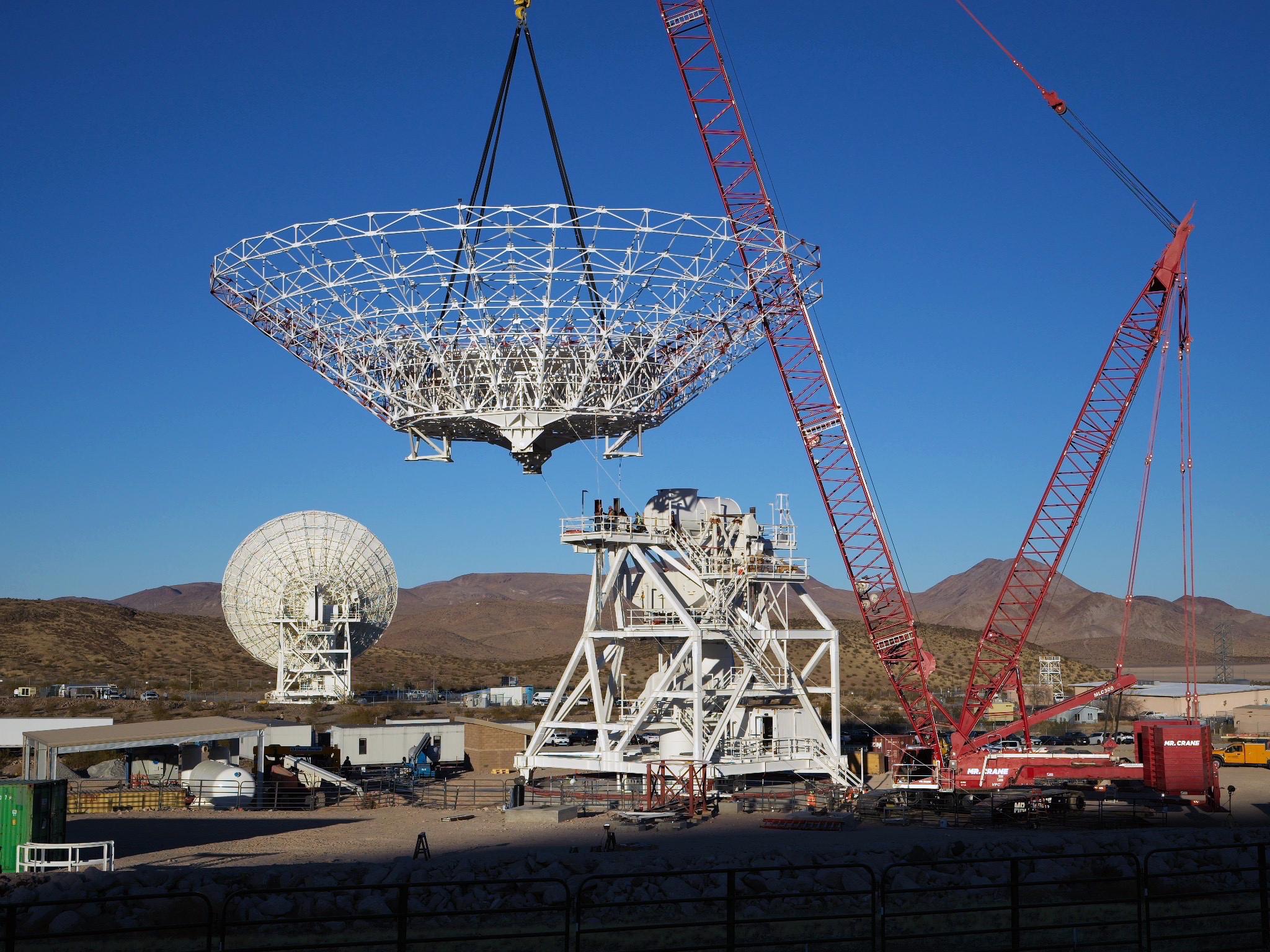 pia26454orig.jpg A large red crane lowers a cone-shaped frame. There is a large white metal structure behind it that has stairs running around it, all the way to the top. In the background at left, a completed antenna faces away from us. Farther away in the background are brown mountains.