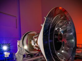 NASA Unveils SPHEREx’s Innovative Concentric Cone Design A spacecraft with a distinct cone shape sits in a clean room. A person in a white suit that covers them from head to toe shines a penlight on the observatory. The walls of the clean room are lit with blue and red lights.