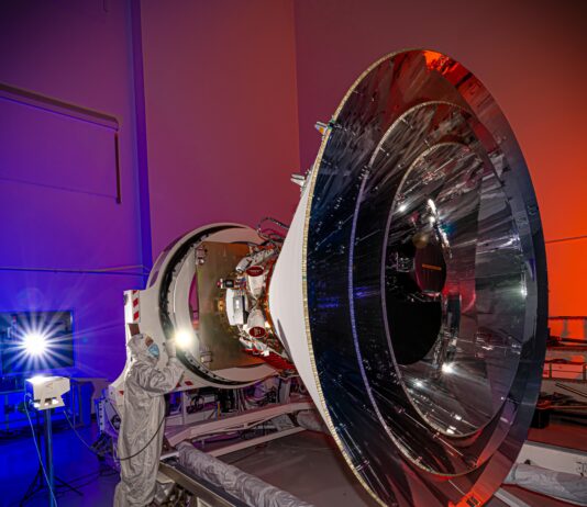 NASA Unveils SPHEREx’s Innovative Concentric Cone Design A spacecraft with a distinct cone shape sits in a clean room. A person in a white suit that covers them from head to toe shines a penlight on the observatory. The walls of the clean room are lit with blue and red lights.