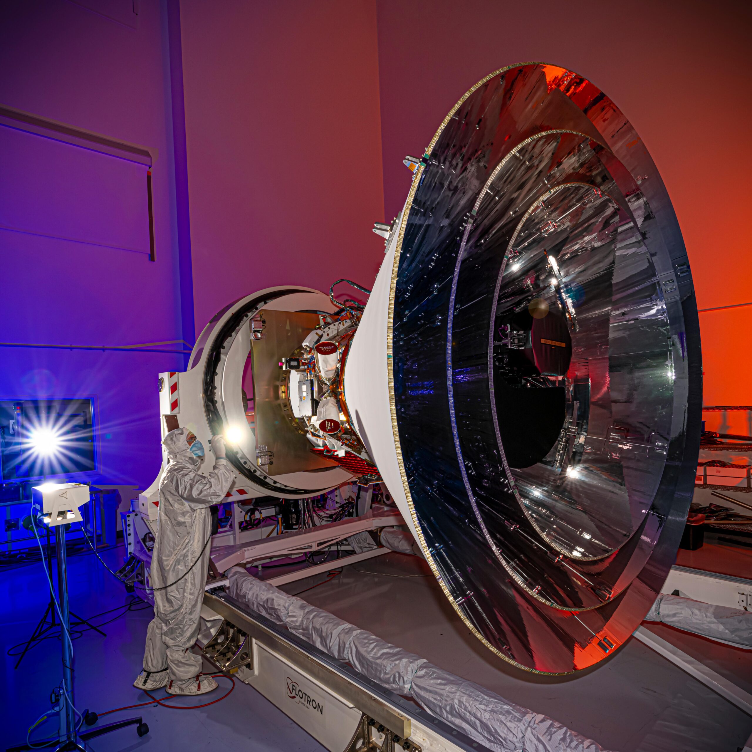 A spacecraft with a distinct cone shape sits in a clean room. A person in a white suit that covers them from head to toe shines a penlight on the observatory. The walls of the clean room are lit with blue and red lights.